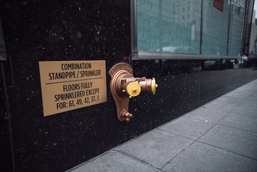 A metallic standpipe and sprinkler connection is mounted on a dark exterior wall, accompanied by a sign indicating the combination standpipe and sprinkler system. The sign mentions that all floors except certain numbered ones are fully sprinklered. The sidewalk next to the wall is tiled, and a large glass window reflects the urban environment.