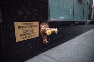 A metallic standpipe and sprinkler connection is mounted on a dark exterior wall, accompanied by a sign indicating the combination standpipe and sprinkler system. The sign mentions that all floors except certain numbered ones are fully sprinklered. The sidewalk next to the wall is tiled, and a large glass window reflects the urban environment.
