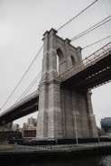 A large stone bridge tower with intricate design and cables extending from the top, situated in an urban area. The surrounding area includes modern buildings and is bordered by a railing alongside a body of water.