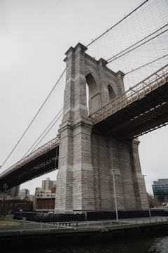 A large stone bridge tower with intricate design and cables extending from the top, situated in an urban area. The surrounding area includes modern buildings and is bordered by a railing alongside a body of water.