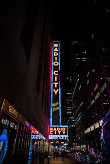 Neon signs brightly illuminate a city street at night, prominently showcasing 'Radio City Music Hall'. Tall buildings flank the signage, with reflective surfaces casting colorful light reflections along the sidewalk. Pedestrians can be seen walking along the dimly lit pavement.