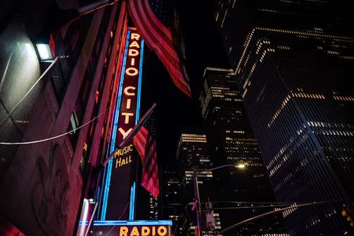 A brightly lit neon sign for Radio City Music Hall stands against a backdrop of towering skyscrapers and an American flag, creating a vibrant nighttime urban scene.