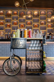 A mobile bar cart is positioned in front of a decorative wall with shelves, featuring a variety of colorful beverage dispensers lined up on top. The wall has a geometric pattern and is adorned with round, vibrant art pieces. Hanging above is a modern light fixture with multiple bulbs, casting a warm glow.