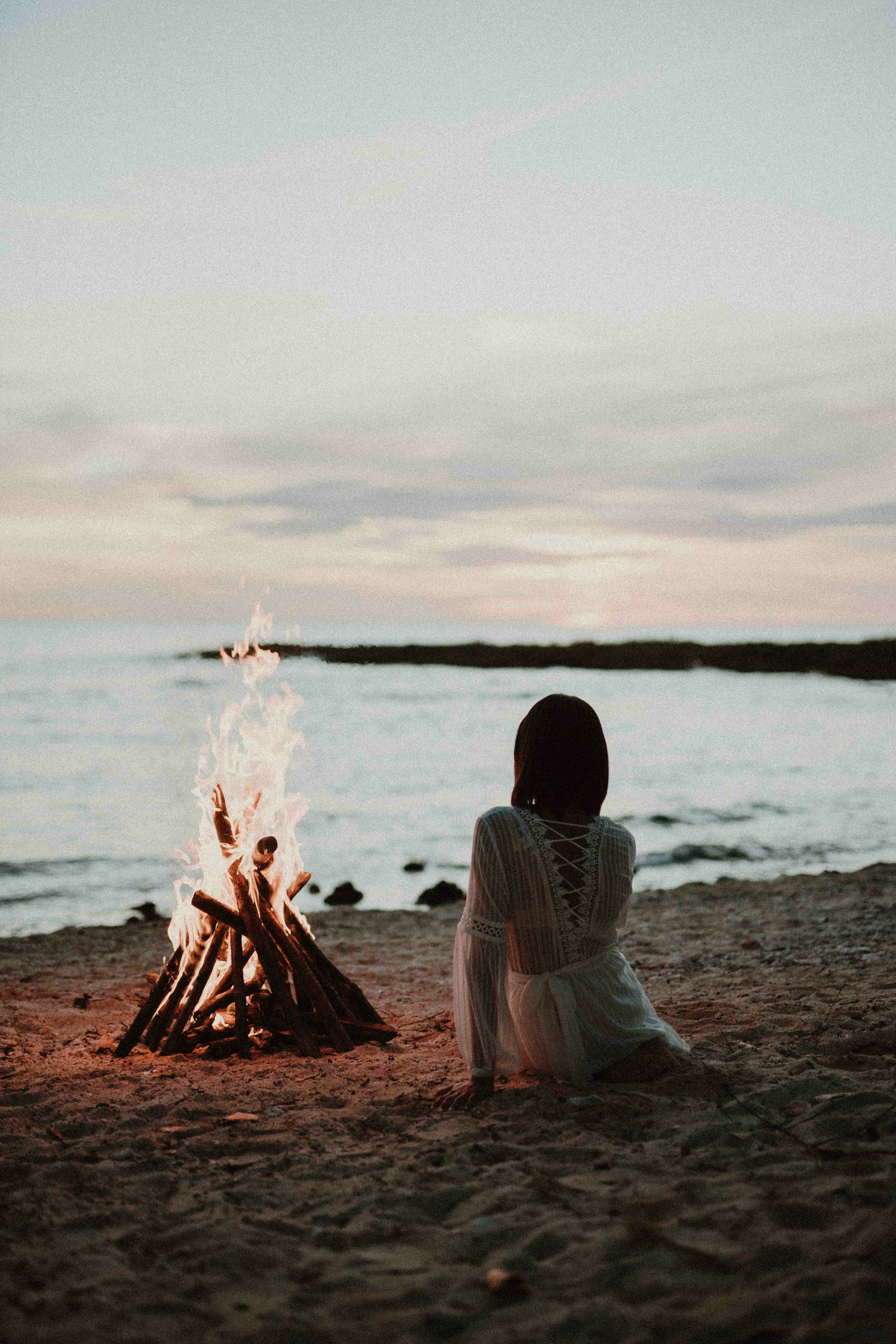 a woman sitting in front of a fire on a beach