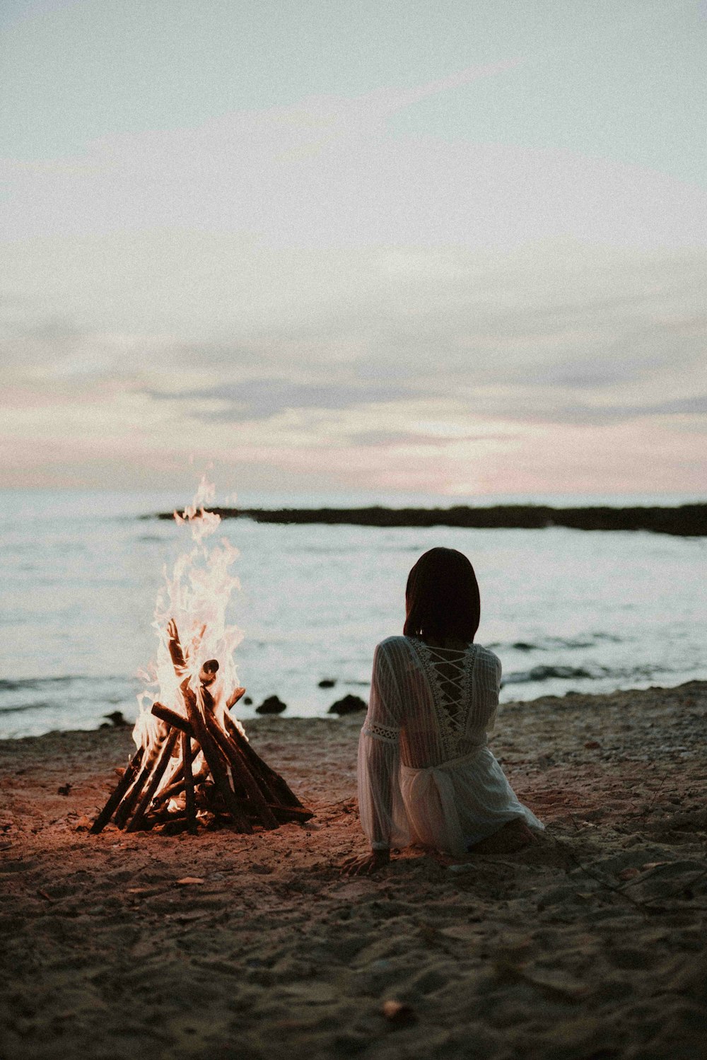 a woman sitting in front of a fire on a beach