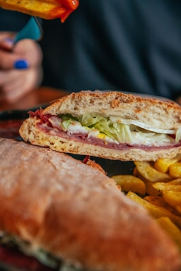 A sandwich with layers of lettuce, deli meat, and cheese inside a sliced baguette. Accompanied by a serving of crispy fries. A hand holding a fry dipped in red sauce is visible in the background.