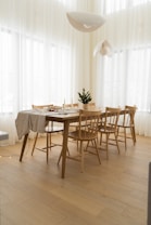 A spacious dining area with a wooden table and chairs set on a light wooden floor. The table is partially covered with a white cloth, and small decorative items are arranged on top, including a miniature Christmas tree and candles. Large pendant lights hang from the ceiling, and sheer curtains allow soft natural light to filter into the room.