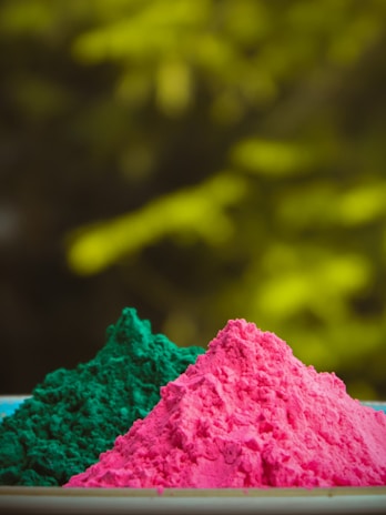 Two vibrant piles of colored powder, one pink and one green, are prominently displayed in the foreground with a soft-focus background of yellowish-green hues.