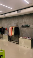 A modern retail clothing store displays a range of colorful hoodies and sweatshirts on a rack against a concrete wall. Below the rack, there are boxes displaying a variety of sneakers. Nearby, other clothing items are neatly folded. The ceiling features sleek, linear lighting and exposed white ductwork.