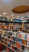 A well-organized bookstore interior with shelves full of diverse books. The sections include categories like Business & Management, Self Help, Indian Fiction, Crime & Mystery, and Bestsellers. The ceiling features recessed lighting and a circular design element.