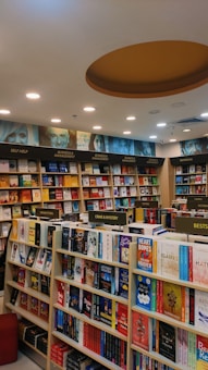 A well-organized bookstore interior with shelves full of diverse books. The sections include categories like Business & Management, Self Help, Indian Fiction, Crime & Mystery, and Bestsellers. The ceiling features recessed lighting and a circular design element.