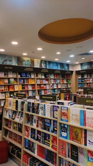 A well-organized bookstore interior with shelves full of diverse books. The sections include categories like Business & Management, Self Help, Indian Fiction, Crime & Mystery, and Bestsellers. The ceiling features recessed lighting and a circular design element.