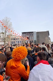 A large crowd of people gathered outdoors, many wearing orange clothing and accessories. Orange and white bunting is strung across the area, and some people are wearing orange wigs. The background features buildings and a clear sky.
