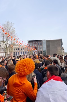 A large crowd of people gathered outdoors, many wearing orange clothing and accessories. Orange and white bunting is strung across the area, and some people are wearing orange wigs. The background features buildings and a clear sky.