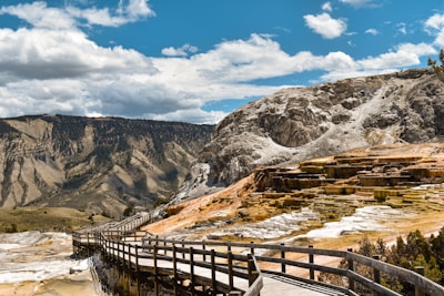 A scenic boardwalk winds through a rocky landscape with layered mineral deposits and terraces. The surrounding mountains stand tall in the background under a bright blue sky filled with fluffy white clouds.