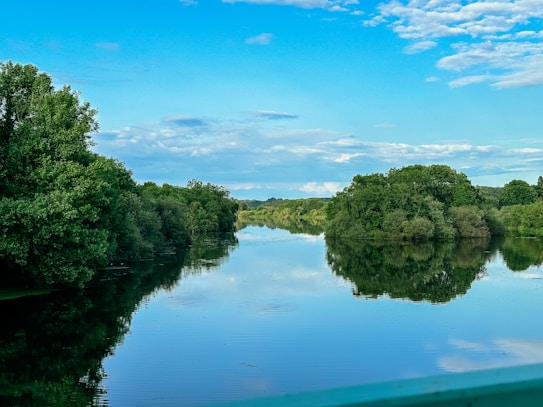A tranquil river reflecting the clear blue sky and surrounding dense green trees. The scene is calm and serene, with fluffy white clouds scattered across the sky.