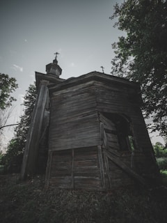 A dilapidated wooden building with religious architecture sits amidst overgrown vegetation. The structure is weathered, with broken panels and a leaning silhouette, crowned with a cross at its peak.