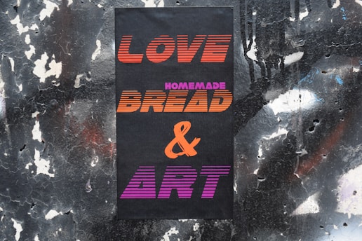 A black poster with vibrant text featuring the words 'LOVE', 'HOMEMADE', 'BREAD', '&', and 'ART'. The background is a rough, textured wall with peeling black and white paint.
