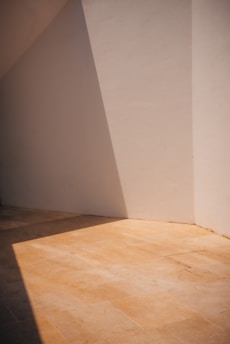 A minimalist scene with a beige wall and floor creating a simple yet striking geometric play of light and shadow. The sunlight casts an angular shadow across the tiled floor, emphasizing the clean lines and serene atmosphere.