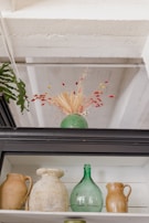 A green vase with intricate patterns contains a dry floral arrangement, featuring red berries and beige botanical elements. Below, various ceramic and glass vessels, including a green glass bottle and brown ceramic jugs, are displayed on a white shelf. A leafy plant partially frames the scene to the left.