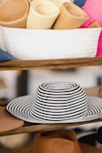 A collection of rolled-up straw hats placed inside a white woven basket sitting on a wooden shelf. Below the basket, a wide-brimmed hat with black and white stripes is prominently displayed, with several other tan-colored hats visible around it.