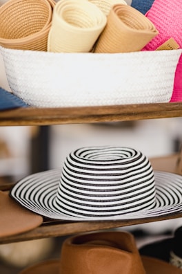 A collection of rolled-up straw hats placed inside a white woven basket sitting on a wooden shelf. Below the basket, a wide-brimmed hat with black and white stripes is prominently displayed, with several other tan-colored hats visible around it.