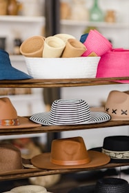 A variety of hats displayed on wooden shelves, including rolled-up hats in a white basket, a striped black and white hat, tan and brown hats, and a pink hat. The background is slightly blurred, giving a focus on the hats.