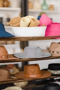 A variety of hats displayed on wooden shelves, including rolled-up hats in a white basket, a striped black and white hat, tan and brown hats, and a pink hat. The background is slightly blurred, giving a focus on the hats.