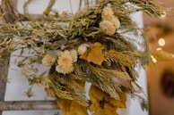 An arrangement of dried yellow flowers and foliage is set against a blurred background, creating a rustic and natural aesthetic. The dried petals and leaves are intricately layered, giving a textured look.
