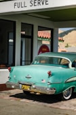 A vintage teal car with a black and yellow California license plate is parked under a 'FULL SERVE' gas station awning. There is an American flag sticker on the rear bumper. The building has white walls with green trim and a red tiled roof. A retro 'Drink Coca-Cola' sign and 'RESTROOM' sign are visible through the glass window.