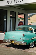 A vintage teal car with a black and yellow California license plate is parked under a 'FULL SERVE' gas station awning. There is an American flag sticker on the rear bumper. The building has white walls with green trim and a red tiled roof. A retro 'Drink Coca-Cola' sign and 'RESTROOM' sign are visible through the glass window.