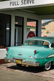A vintage teal car with a black and yellow California license plate is parked under a 'FULL SERVE' gas station awning. There is an American flag sticker on the rear bumper. The building has white walls with green trim and a red tiled roof. A retro 'Drink Coca-Cola' sign and 'RESTROOM' sign are visible through the glass window.
