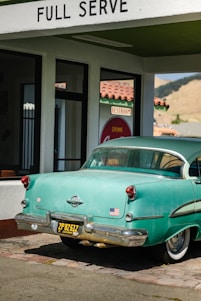 A vintage teal car with a black and yellow California license plate is parked under a 'FULL SERVE' gas station awning. There is an American flag sticker on the rear bumper. The building has white walls with green trim and a red tiled roof. A retro 'Drink Coca-Cola' sign and 'RESTROOM' sign are visible through the glass window.