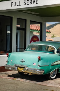 A vintage teal car with a black and yellow California license plate is parked under a 'FULL SERVE' gas station awning. There is an American flag sticker on the rear bumper. The building has white walls with green trim and a red tiled roof. A retro 'Drink Coca-Cola' sign and 'RESTROOM' sign are visible through the glass window.