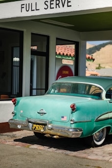 A vintage teal car with a black and yellow California license plate is parked under a 'FULL SERVE' gas station awning. There is an American flag sticker on the rear bumper. The building has white walls with green trim and a red tiled roof. A retro 'Drink Coca-Cola' sign and 'RESTROOM' sign are visible through the glass window.