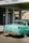 A vintage teal car with a black and yellow California license plate is parked under a 'FULL SERVE' gas station awning. There is an American flag sticker on the rear bumper. The building has white walls with green trim and a red tiled roof. A retro 'Drink Coca-Cola' sign and 'RESTROOM' sign are visible through the glass window.