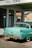 A vintage teal car with a black and yellow California license plate is parked under a 'FULL SERVE' gas station awning. There is an American flag sticker on the rear bumper. The building has white walls with green trim and a red tiled roof. A retro 'Drink Coca-Cola' sign and 'RESTROOM' sign are visible through the glass window.