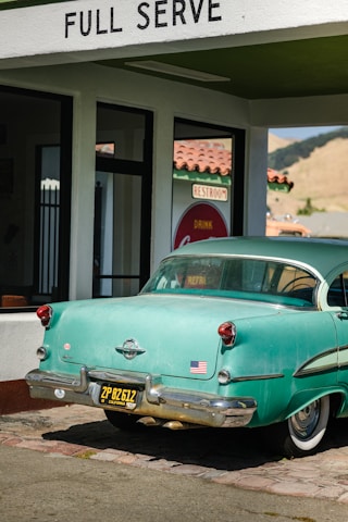 A vintage teal car with a black and yellow California license plate is parked under a 'FULL SERVE' gas station awning. There is an American flag sticker on the rear bumper. The building has white walls with green trim and a red tiled roof. A retro 'Drink Coca-Cola' sign and 'RESTROOM' sign are visible through the glass window.