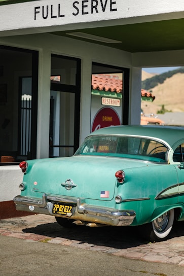A vintage teal car with a black and yellow California license plate is parked under a 'FULL SERVE' gas station awning. There is an American flag sticker on the rear bumper. The building has white walls with green trim and a red tiled roof. A retro 'Drink Coca-Cola' sign and 'RESTROOM' sign are visible through the glass window.