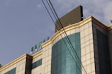 The image depicts an upper section of a building with a modern architectural design. It features a facade made of reflective glass panels, outlined with golden-colored framing. A visible sign on the building reads 'DAMAI HOTEL' in teal letters. Overhead, electrical wires intersect the view against a blue sky backdrop.