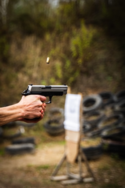 A person's hand is firing a handgun, with the bullet casing visible in mid-air. The setting appears to be an outdoor shooting range, with tires and a target stand blurred in the background.