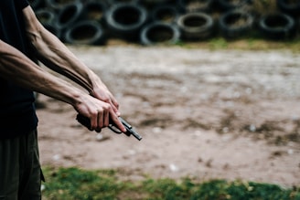 A close-up of a person holding a handgun with both hands, with visible muscles and veins on the arms. In the background, there is a stack of old tires arranged in rows. The scene appears to be outdoors on a dirt or gravel surface, with grass visible in the foreground.