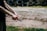 A close-up of a person holding a handgun with both hands, with visible muscles and veins on the arms. In the background, there is a stack of old tires arranged in rows. The scene appears to be outdoors on a dirt or gravel surface, with grass visible in the foreground.