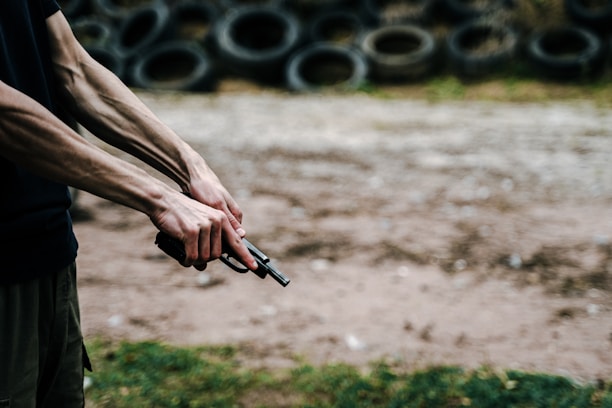 A close-up of a person holding a handgun with both hands, with visible muscles and veins on the arms. In the background, there is a stack of old tires arranged in rows. The scene appears to be outdoors on a dirt or gravel surface, with grass visible in the foreground.