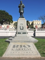 A statue of a historical figure stands on a tall pedestal in a park setting. The base of the statue is engraved with 'PII' and laurel designs. In the foreground, there is a tiled pavement with the words 'RIO CAPITAL DA INDEPENDENCIA 1822-2022' surrounded by green grass. In the background, an elegant building with neoclassical architecture is visible under a clear blue sky.