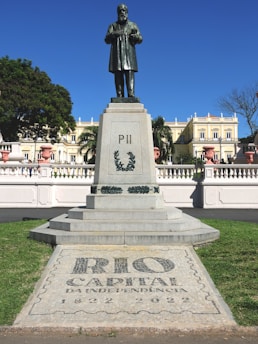 A statue of a historical figure stands on a tall pedestal in a park setting. The base of the statue is engraved with 'PII' and laurel designs. In the foreground, there is a tiled pavement with the words 'RIO CAPITAL DA INDEPENDENCIA 1822-2022' surrounded by green grass. In the background, an elegant building with neoclassical architecture is visible under a clear blue sky.