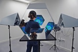 A person stands in a room with advanced lighting equipment. They are wearing headphones and their face is not visible. Mirrors and multiple hexagonal structures with glowing blue lights are arranged around them, creating a futuristic and reflective environment. The walls are white and there are tripod stands supporting the mirrors.