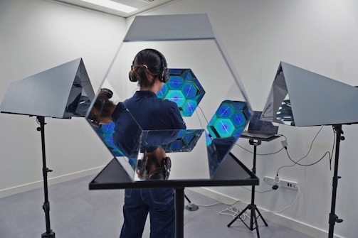 A person stands in a room with advanced lighting equipment. They are wearing headphones and their face is not visible. Mirrors and multiple hexagonal structures with glowing blue lights are arranged around them, creating a futuristic and reflective environment. The walls are white and there are tripod stands supporting the mirrors.