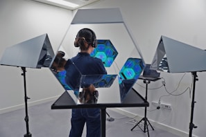 A person stands in a room with advanced lighting equipment. They are wearing headphones and their face is not visible. Mirrors and multiple hexagonal structures with glowing blue lights are arranged around them, creating a futuristic and reflective environment. The walls are white and there are tripod stands supporting the mirrors.