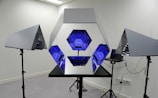 A hexagonal mirror structure is flanked by two triangular mirrors on stands, with blue geometric patterns inside. The setup is in a room with a laptop connected to the mirrors.